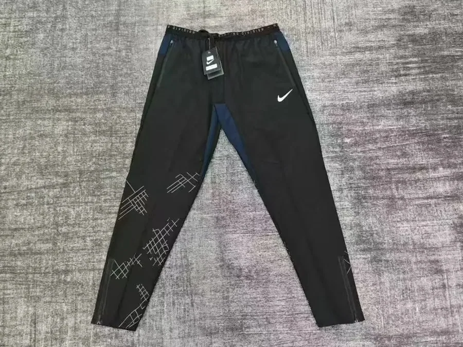 Pantalón de chándal negro con detalles reflectantes colgado de una percha sobre una pared blanca